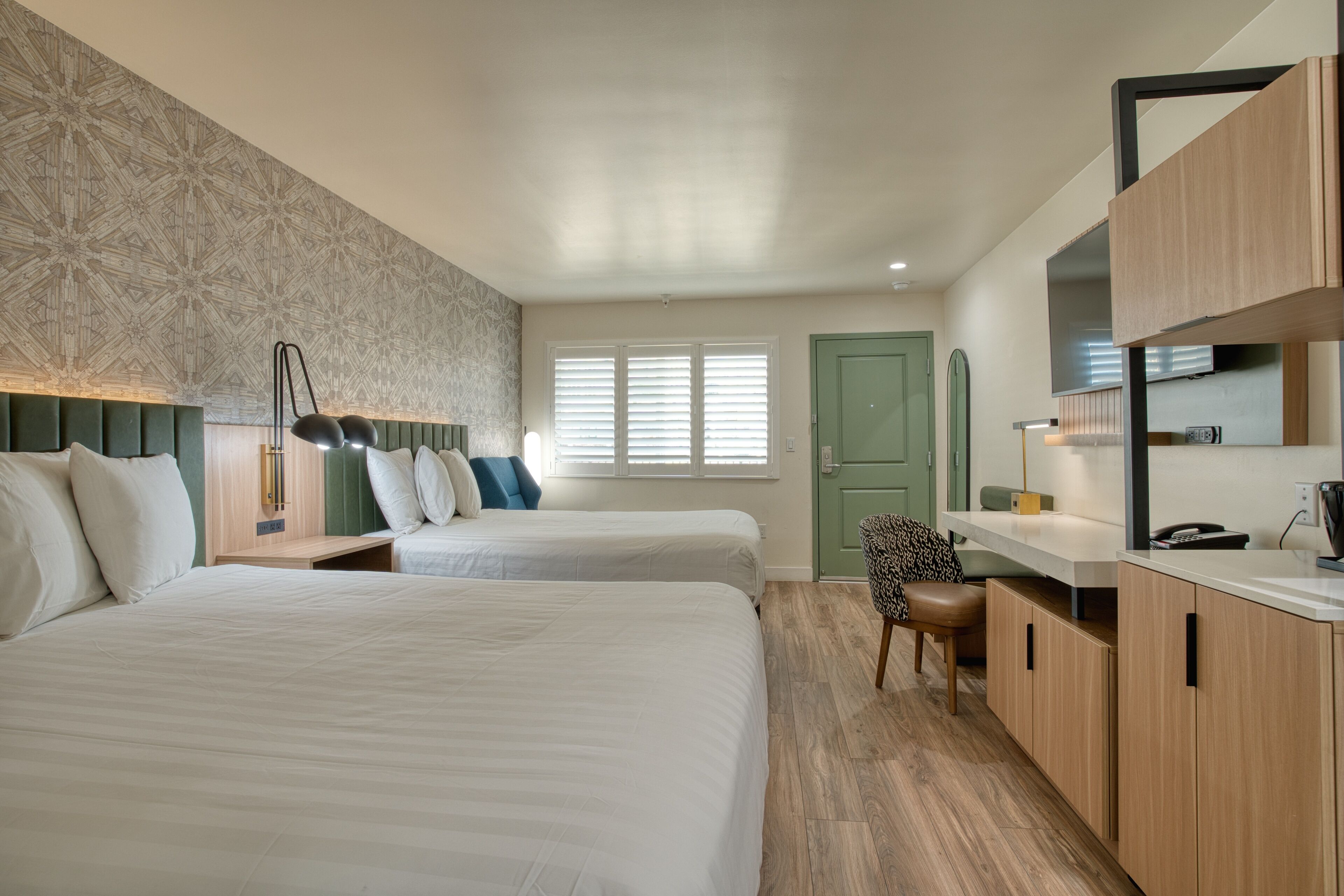 A hotel room with two beds, a desk, a chair, and a green door.