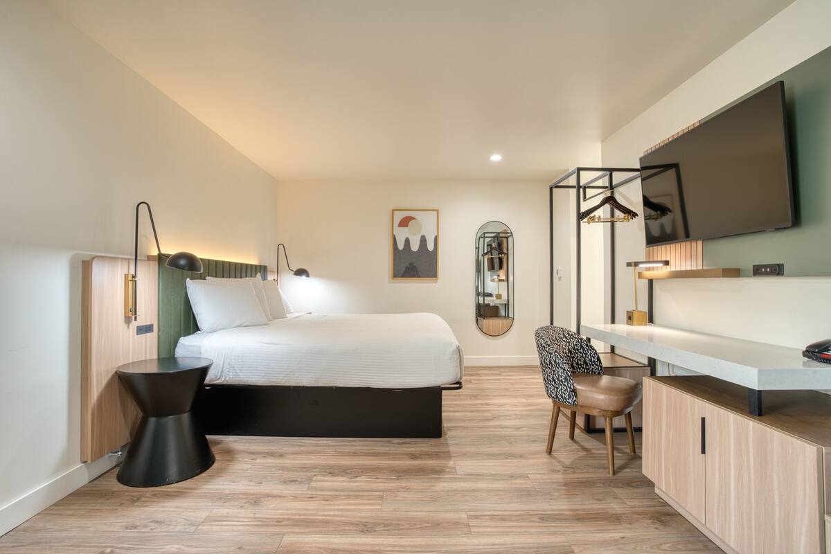 Una habitación de hotel moderna con cama, mesita de noche, silla, escritorio y un espejo grande.