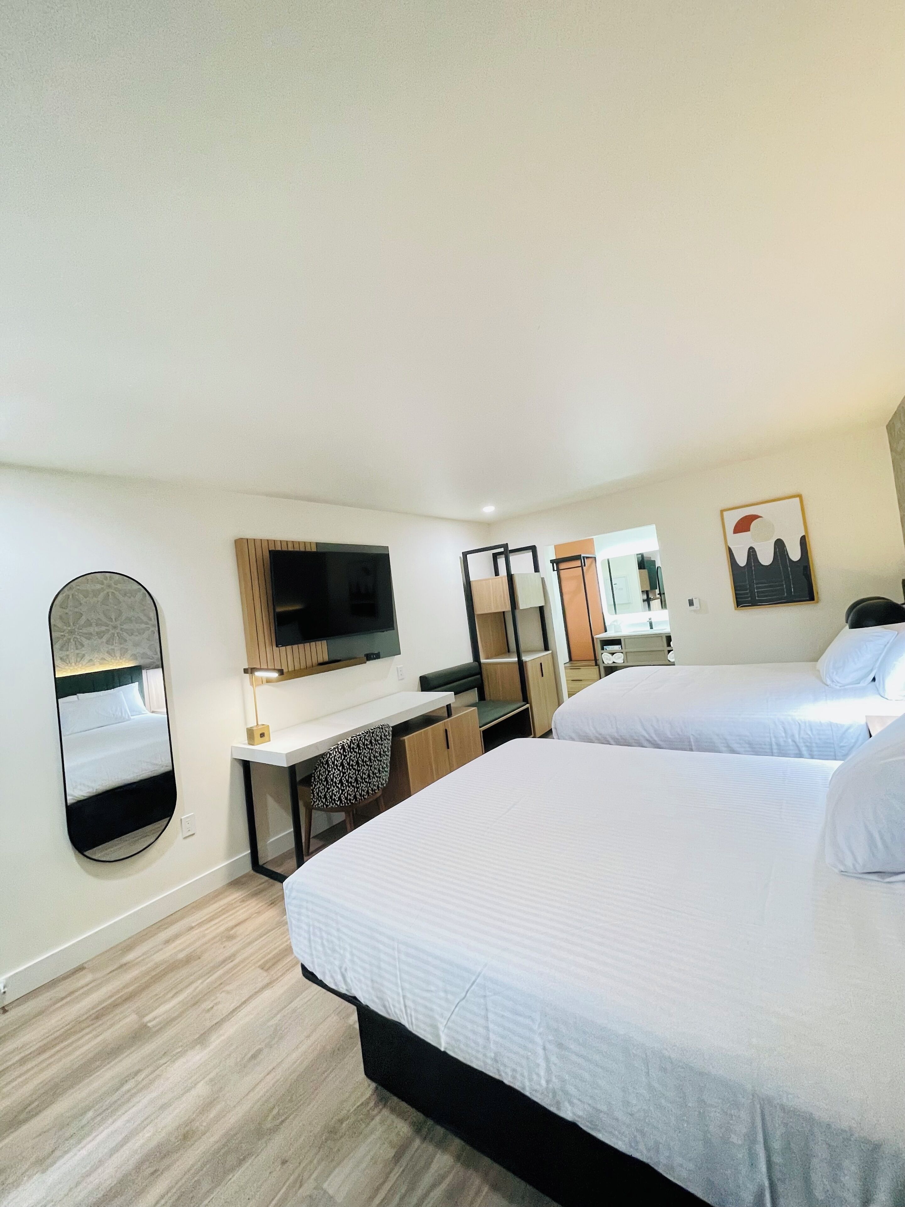 Habitación de hotel con una cama grande, un escritorio con silla, un televisor, un espejo y un ventanal pequeño.