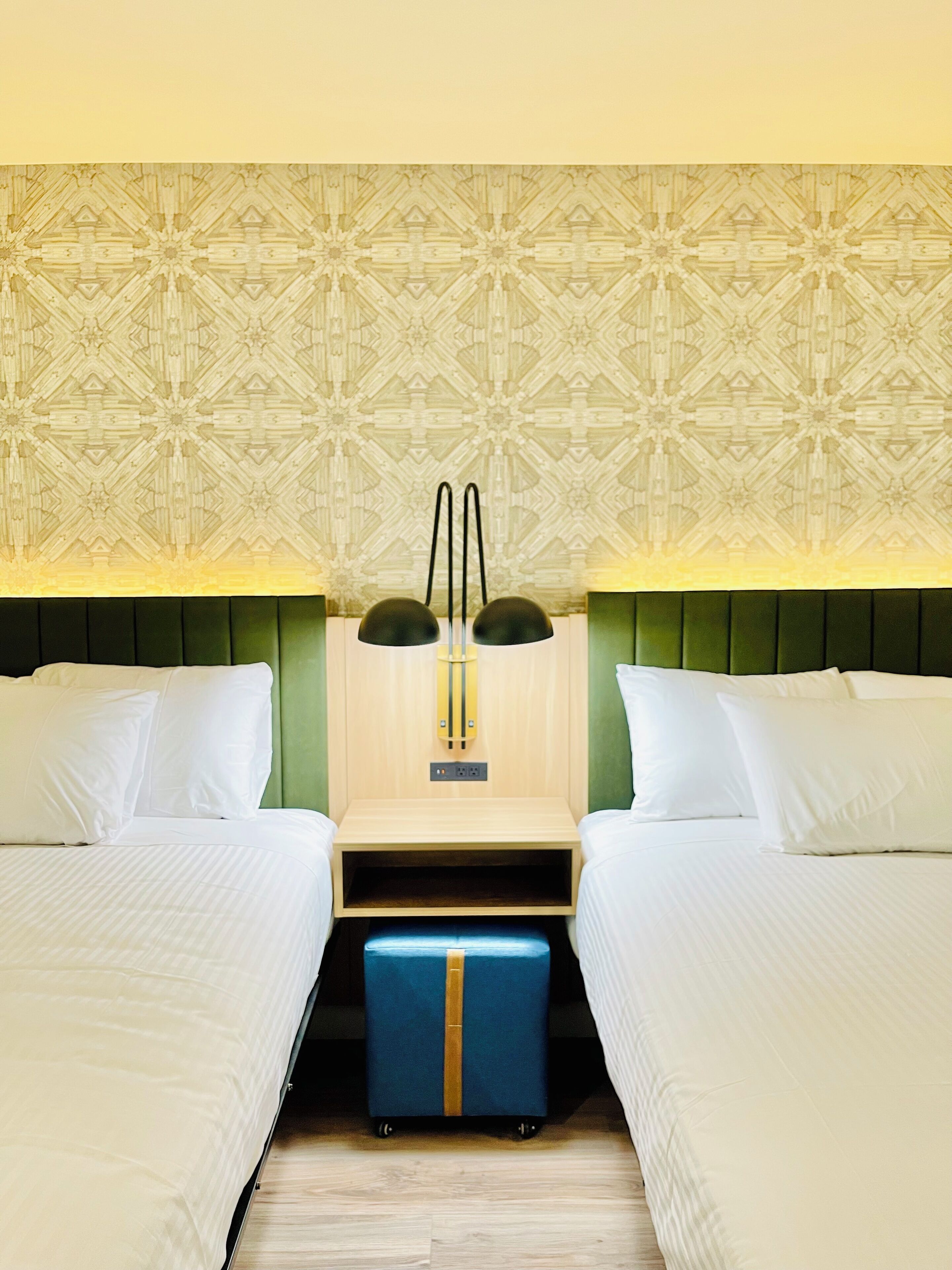 A hotel room with two beds, a patterned wall, a headboard, and a small table with a suitcase.