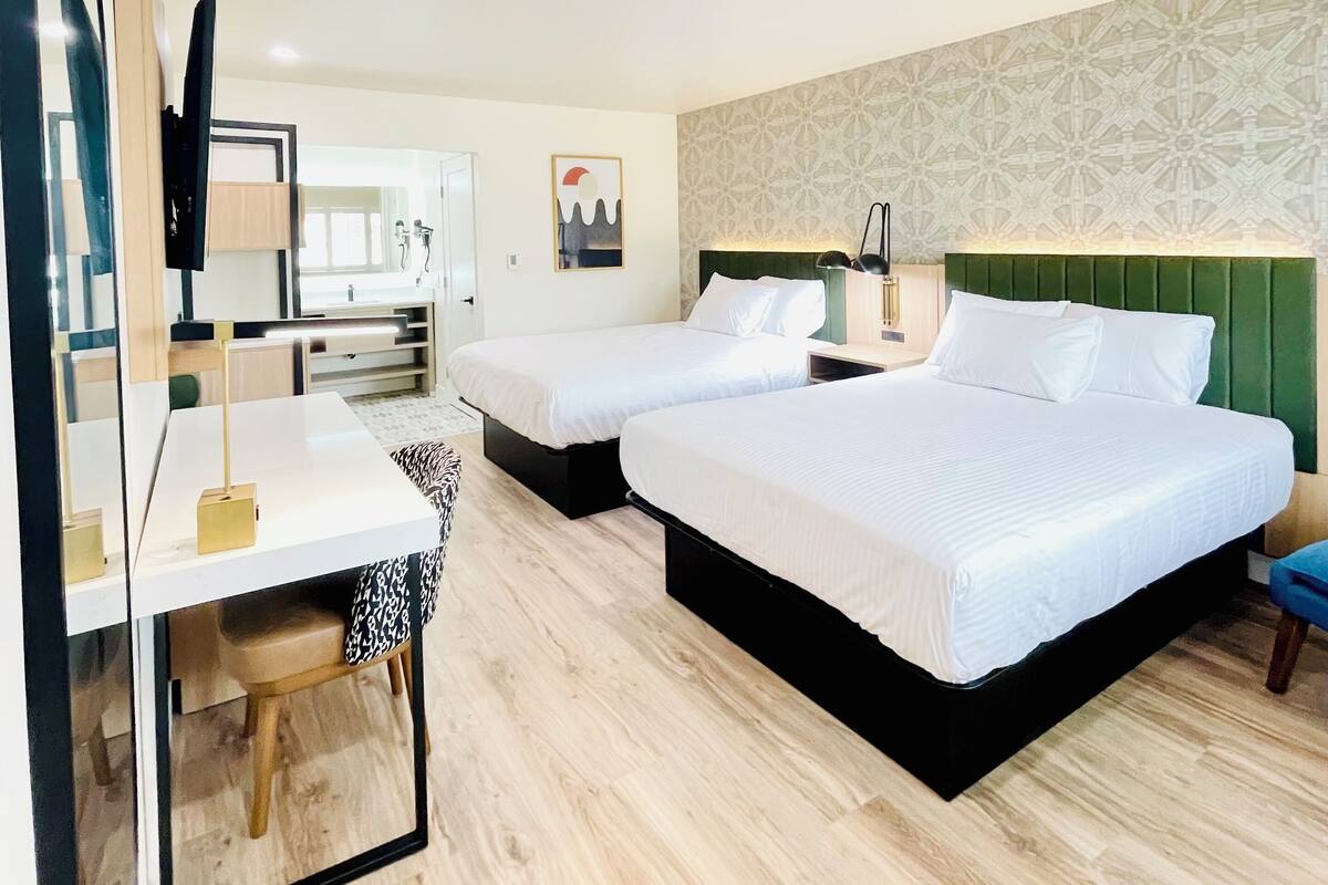 Habitación de hotel con dos camas, un escritorio, una silla, un espejo y un cabecero verde.