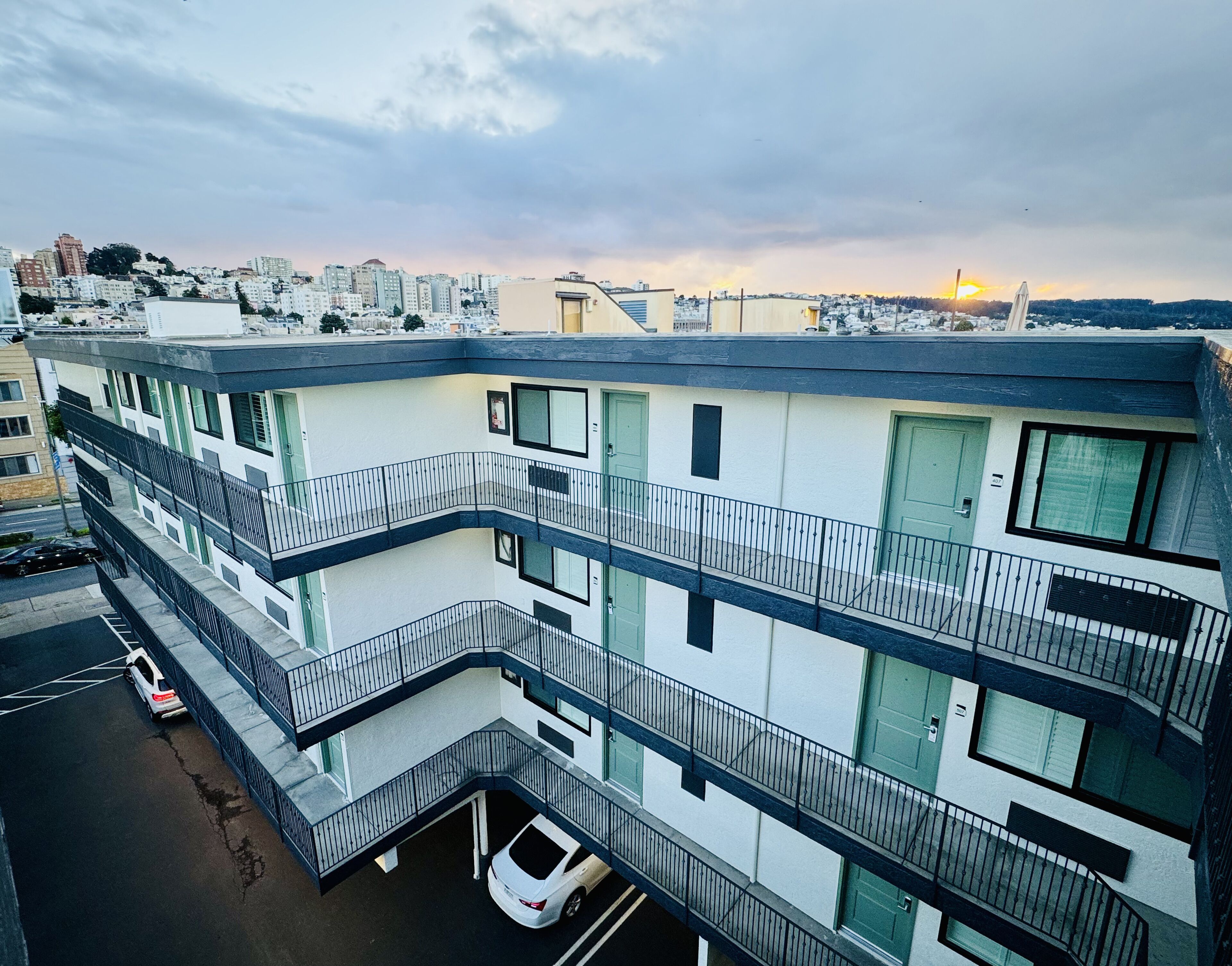 A multi-story residential building with balconies, a parking area, and a view of the cityscape in the background.
