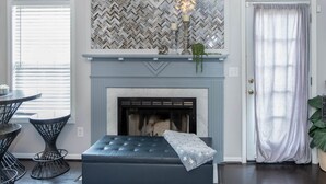 A living room with a fireplace, a dining table with chairs, and a large window with blinds. - The Herringbone Blue (Harnett County)