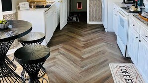 A compact kitchen with white cabinets, a herringbone wood floor, and a coffee-themed rug. - The Herringbone Blue (Harnett County)