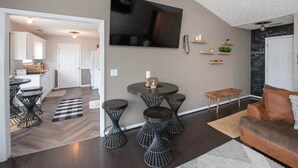 A modern living room with a round coffee table, four black stools, a brown sofa, and a flat-screen TV mounted on the wall. - The Herringbone Blue (Harnett County)