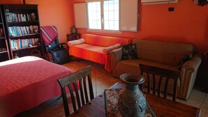 Une pièce confortable avec un canapé, un fauteuil, une petite table sur laquelle est posé un vase, une bibliothèque et une fenêtre avec des stores.