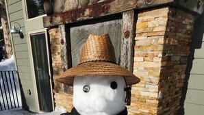 Un muñeco de nieve con sombrero de paja y bufanda frente a un muro de ladrillo.