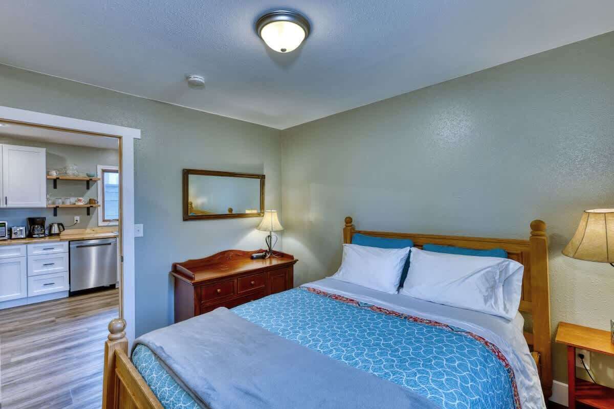 A hotel room with a bed, bedside table, lamp, and a view of the kitchen area.