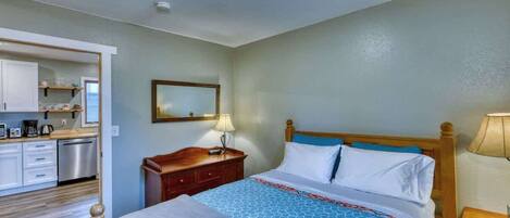 A hotel room with a bed, bedside table, lamp, and a view of the kitchen area.