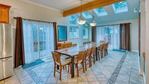 A spacious kitchen with a long dining table, wooden cabinets, and a view through large windows. - Grandeur Gathering Luxurious retreat with hot tub (Kodiak)