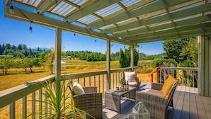 A covered outdoor seating area with wicker furniture and a glass enclosure, overlooking a field. - Combined Sequim · Spacious Farmhouse & Vintage Trailer on 5 Acres (Sequim)
