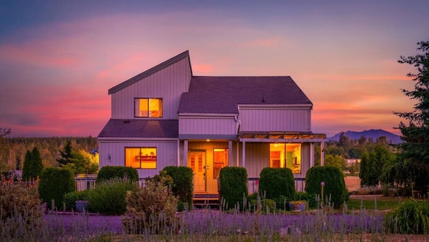 A two-story house with a porch, surrounded by a garden with lavender. - Combined Sequim · Spacious Farmhouse & Vintage Trailer on 5 Acres (Sequim)