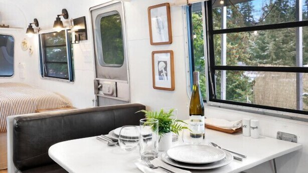 A compact kitchen area with a dining table set for two, a bench, and a window with a view of greenery. - Fungalow · The Fungalow: Vintage Trailer with Modern Comfort (Sequim)