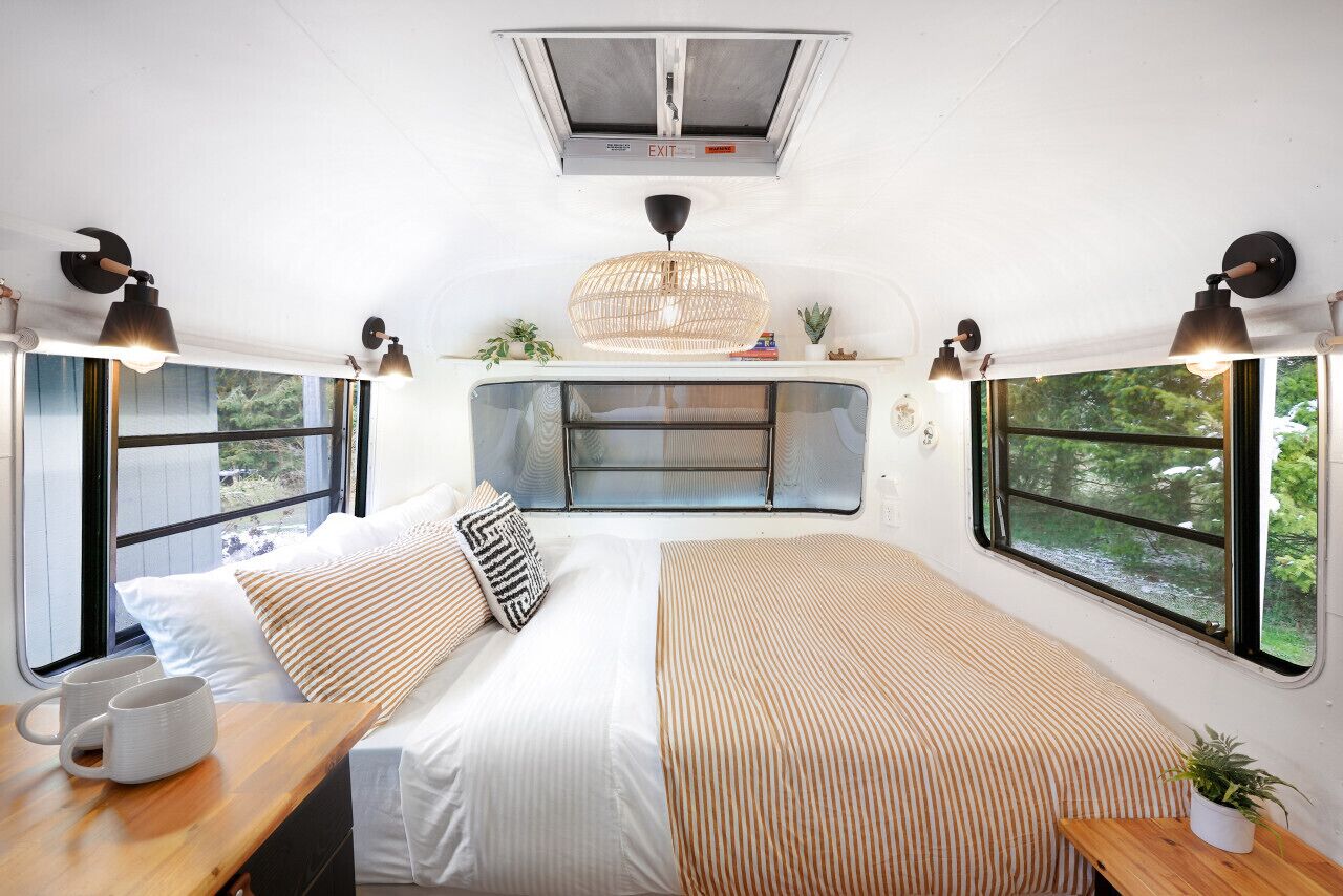 A cozy interior with a bed, bedside table, and two windows offering a view of greenery.