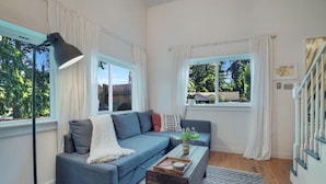 A modern living room with a grey sofa, a small wooden coffee table, and a floor lamp. There are two windows with white curtains, and a staircase is visible to the right. - Modern, Cozy Urban Homestead w/ Loft (Shoreline)
