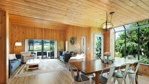 A spacious living area with a wooden ceiling, a dining table with chairs, and a large window providing a view of greenery outside. - Farmhouse · Peaceful Farmhouse with Mountain Views & Hot Tub (Sequim)