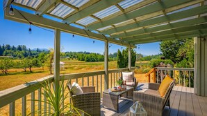 A covered outdoor seating area with wicker furniture and a glass railing overlooking a vineyard. - Farmhouse · Peaceful Farmhouse with Mountain Views & Hot Tub (Sequim)