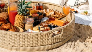 Un montaje para picnic en la playa con una cesta de mimbre llena de comida y bebidas, una piña y una botella de jugo, sobre arena con el océano al fondo.