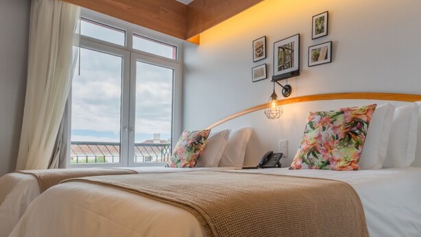 Et hotelværelse med et stort vindue, en seng med blomsterpuder, en sengetavsalampe og en telefon på sengebordet.