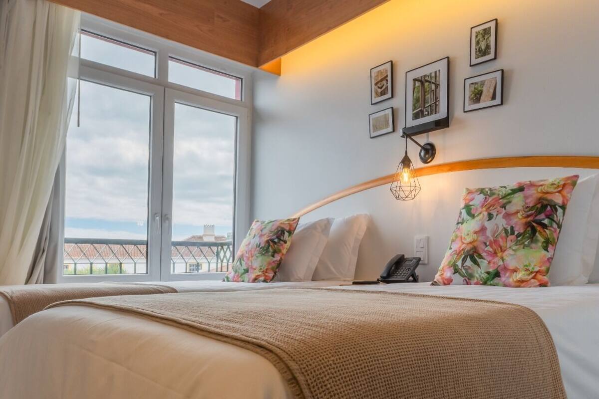 A hotel room with a large window, a bed with floral pillows, a bedside lamp, and a phone on the nightstand.