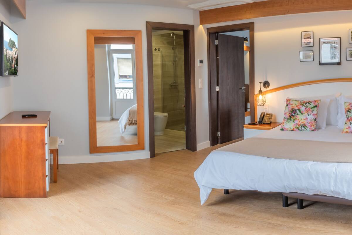 A bedroom with a large bed, a wooden dresser, a mirror, a shower, and a TV on the wall.