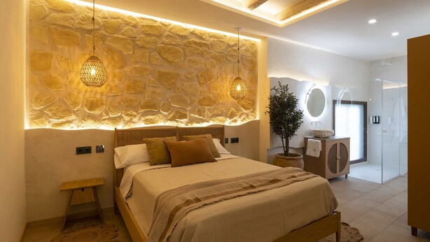 A modern bedroom with a stone wall, a bed with a wooden headboard, two pendant lights, and a large mirror. - Alpujarras | Palatine Concept Suites | Luxury Apartments in Granada Center (Granada)
