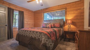 A bedroom with a wooden floor, a bed with patterned bedding, a ceiling fan, a window with blinds, and a bedside lamp.