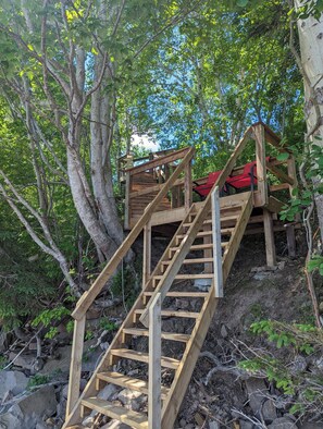A wooden staircase leading to a red bench in a forested area. - 2-Bedroom Cottage On Lake Ainlsie Private Beach, Enjoy Fishing & Swimming. (INVERNESS)