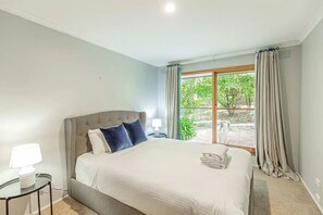A bedroom with a large bed, a bedside table with a lamp, and a view of greenery outside.