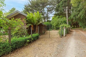 A wooden cabin with a gravel driveway leading to a metal gate, surrounded by dense greenery and trees. - Tranquil 4 Bedroom Cottage in Olinda (Olinda)