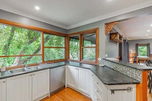 Una cocina con armarios blancos, encimera negra y grandes ventanales con vistas a la vegetación.
