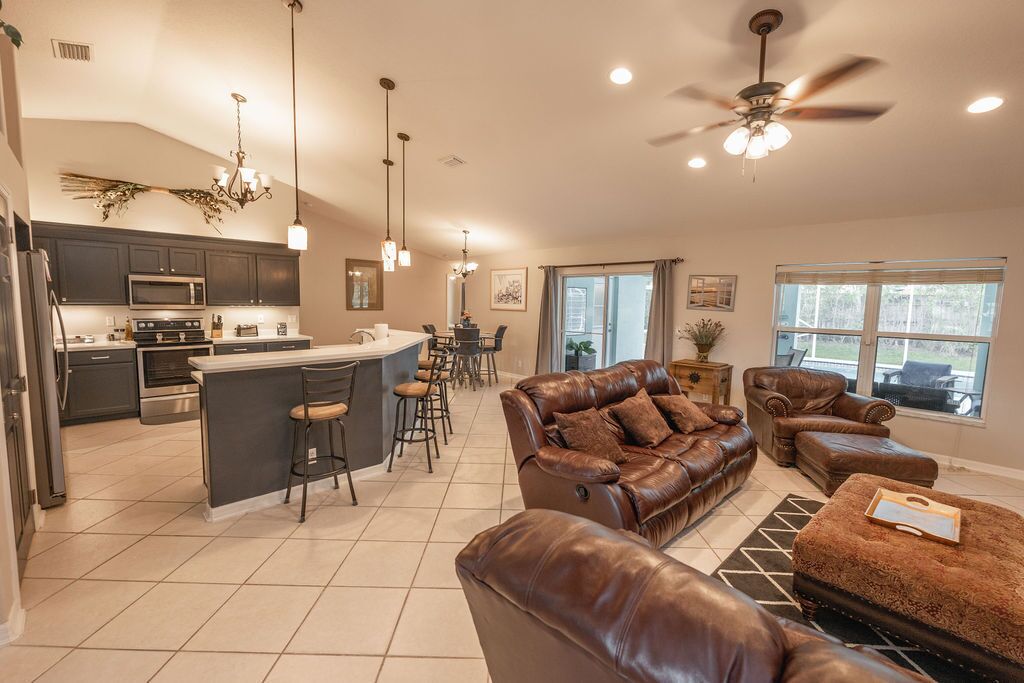 A spacious kitchen with a central island, a dining area with a chandelier, a leather sofa, and a ceiling fan.