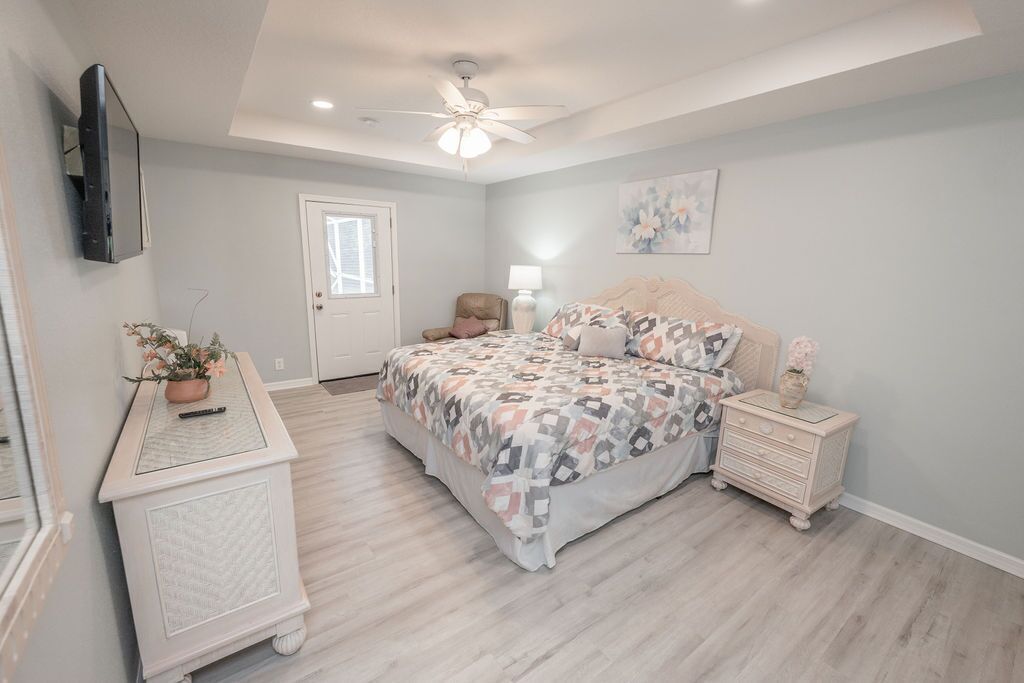 A bedroom with a bed, a nightstand, a dresser, a ceiling fan, a door, and a wall-mounted TV.