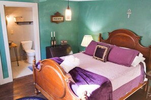 Un dormitorio con cama de madera, ropa de cama morada, una mesita de noche con lámpara y un estante de pared con libros.