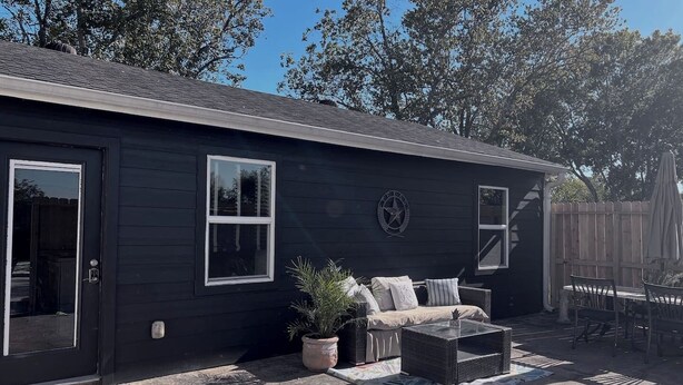 A dark-colored single-story house with a front door, two windows, a porch with a wicker sofa, a small table, and a potted plant. - Quiet, Cozy Guesthouse with privacy (Manvel)