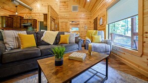 A log cabin interior with a living room featuring a dark leather sofa, a wooden coffee table, and a large window with a view of trees. - 1 BR in Heart of Hochatown - 2 Min Walk to Putt-Putt (Broken Bow)