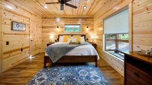 A wooden cabin bedroom with a bed, a ceiling fan, a window, and a nightstand. - 1 BR in Heart of Hochatown - 2 Min Walk to Putt-Putt (Broken Bow)