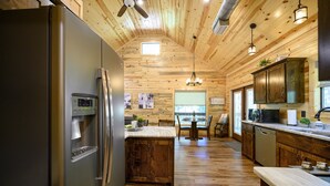 A kitchen with wooden ceiling, stainless steel refrigerator, wooden cabinets, and a dining area with a table and chairs. - 1 BR in Heart of Hochatown - 2 Min Walk to Putt-Putt (Broken Bow)