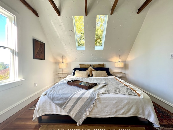 A bedroom with a sloped ceiling, a bed with multiple pillows, two bedside lamps, and a window with a view of trees.