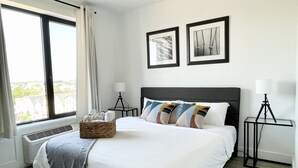 Un dormitorio con una cama grande, una ventana con cortinas, una lámpara de pie y dos cuadros en la pared.