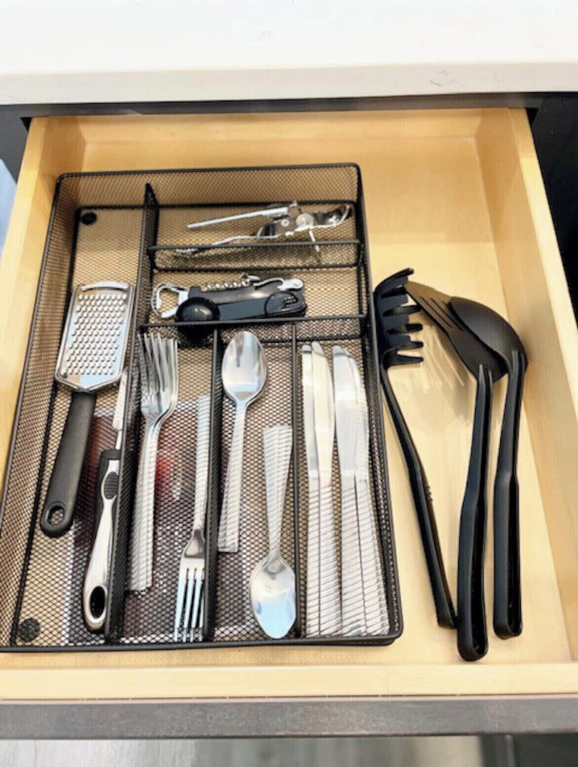 Un cajón con un organizador de malla que contiene diversos utensilios de cocina, como un rallador, unas pinzas, un juego de cuchillos, una cuchara y un tenedor.