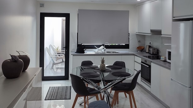 Uma cozinha moderna com armários brancos, um espaço de jantar com mesa de vidro e cadeiras pretas, e uma janela grande com vista para uma varanda.