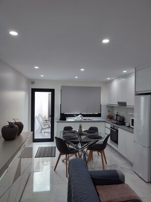 Uma cozinha moderna com armários brancos, um espaço de jantar com mesa de vidro e cadeiras pretas, e uma janela grande com vista para uma varanda.