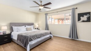 Un dormitorio con cama, mesitas de noche, ventilador de techo, ventana con cortinas y una obra de arte colgada en la pared.