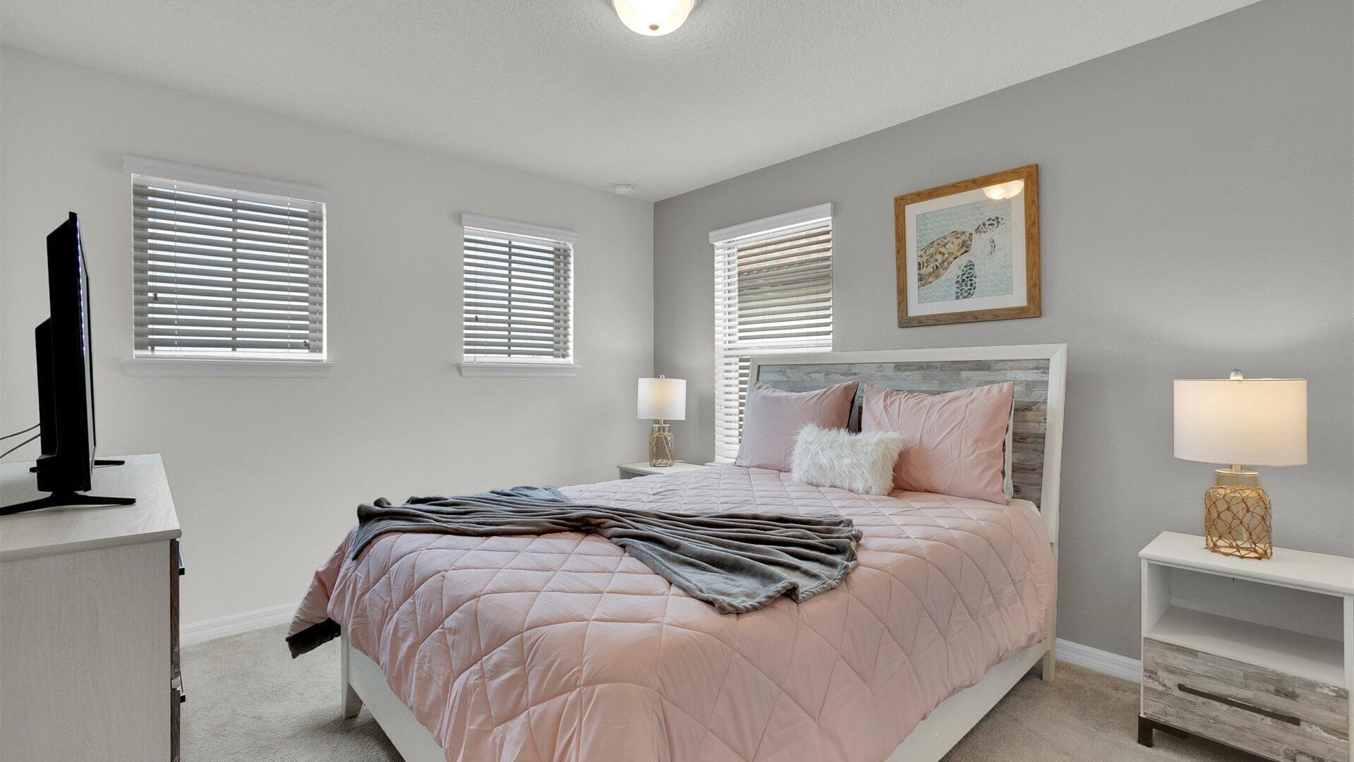 A bedroom with a bed, two bedside tables, a TV, and three windows with blinds.