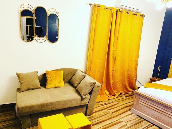 Une pièce comprenant un canapé, deux coussins jaunes, un lit avec une tête de lit en bois, un miroir et des rideaux jaunes.