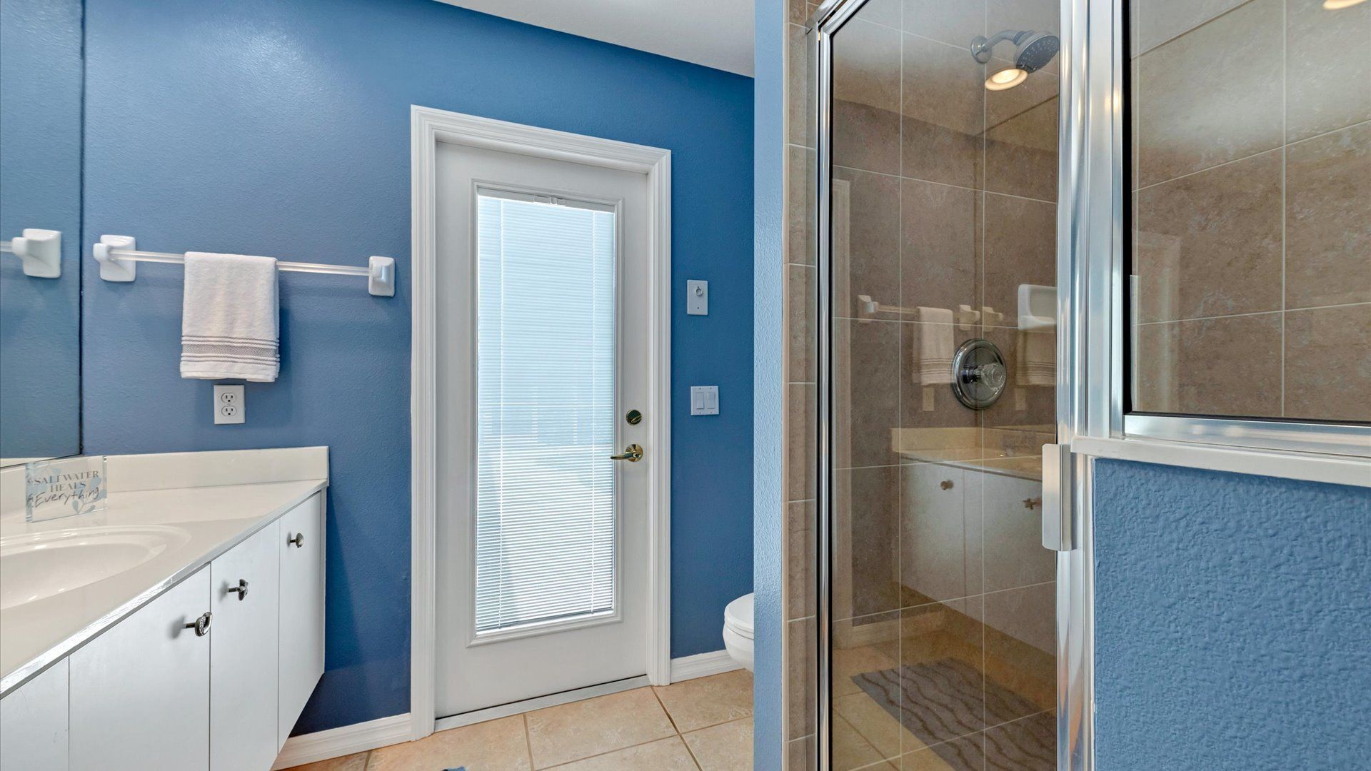 Een badkamer met een blauwe wand, een witte deur, een glazen douchecabine en een wit wastafelmeubel met een handdoekrek.