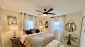 A bedroom with a bed, bedside lamps, a ceiling fan, a window with curtains, a mirror, and a potted plant. - Ocean Beach Cottage (Palm Coast)