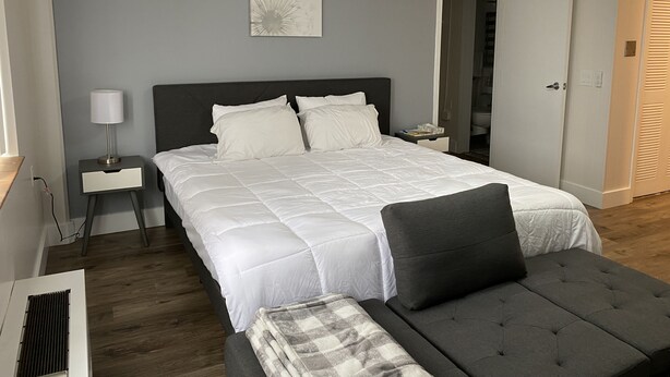 A neatly arranged bedroom with a large bed, bedside tables, a bedside lamp, a gray sofa, and a door leading to another room. - Modernized Studio Apartment in Gadsden, AL (Gadsden)
