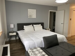 A neatly arranged bedroom with a large bed, bedside tables, a bedside lamp, a gray sofa, and a door leading to another room. - Modernized Studio Apartment in Gadsden, AL (Gadsden)
